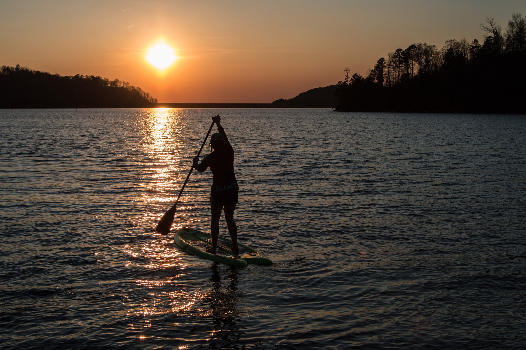 A stand-up paddleboarder stands upright on their blow-up paddleboard, slightly deflated, floating on the gentle waves of Carters Lake in Ellijay, Georgia. They have a paddle in their hands and are actively paddling. In the background are a few shorelines lined with tall trees, and in the sky before the paddleboarder is the setting sun, which paints the entire scene in a golden glow and reflects its shine off the water.